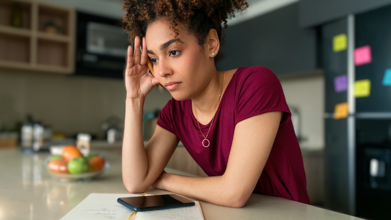 Woman in kitchen, leaning on counter with phone and notebook in front of her. Her head is in her hand, and she is staring into space, feeling overwhelmed | Regain Control Today: 8 Time Management Tips for People with OCD | Mindful Health Solutions