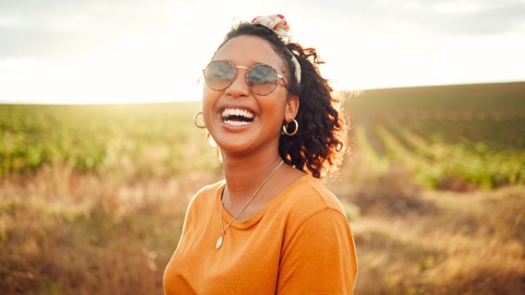 A woman of color outside in a field wearing an orange shirt and sunglasses smiling | The RIse of Esketamine: 7 Reasons Why It's Becoming an Antidepressant Alternative | Mindful Health Solutions