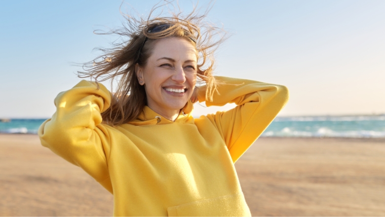Caucasian woman on beach, hair being blown by wind, with a big smile on her face | How To Say Goodbye to Depression FAST Introducing Accelerated TMS | Mindful Health Solutions