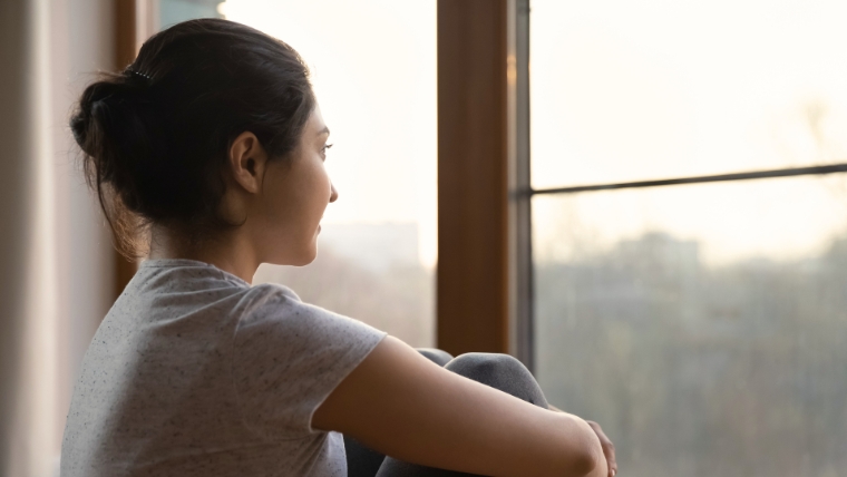 Woman hugging her knees while looking out the window with a hopeful look on her face | Could TMS Help You Explore This Science-Backed Revolution in Mental Health | Mindful Health Solutions