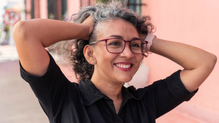 Mature woman with glasses smiling in front a pink wall with her hands in her hair | TMS Therapy Treatment for Chronic Pain Relief: Is It Right for You? | Mindful Health Solutions